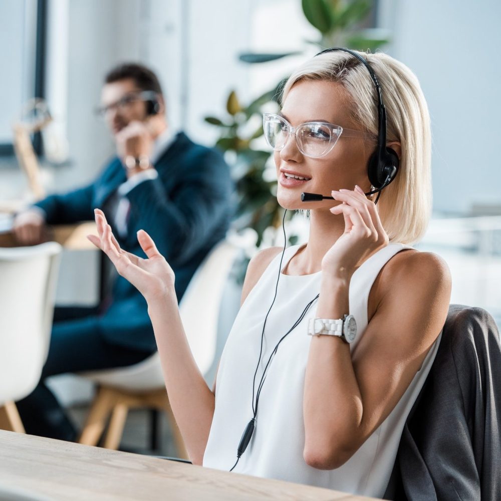 selective-focus-of-happy-operator-touching-headset-talking-and-gesturing-near-coworker-e1640059313540